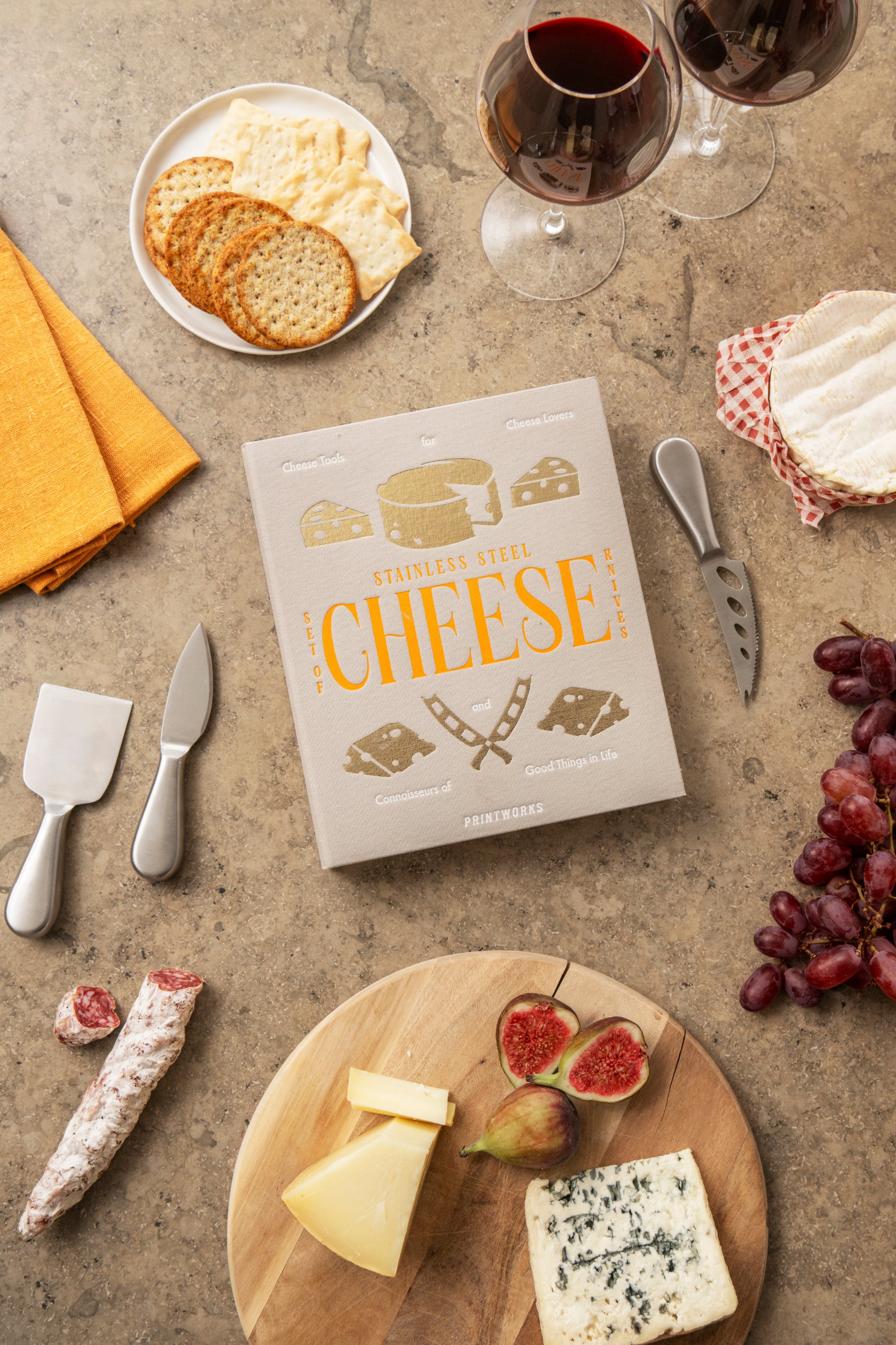 Cheese platter with a book titled 'Mastering Cheese' on a rustic surface with wine, crackers, and figs.